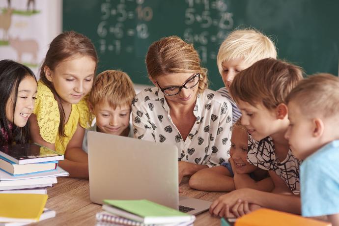 Nauczycielka siedząca z szóstką uczniami przed laptopem w sali lekcyjnej
