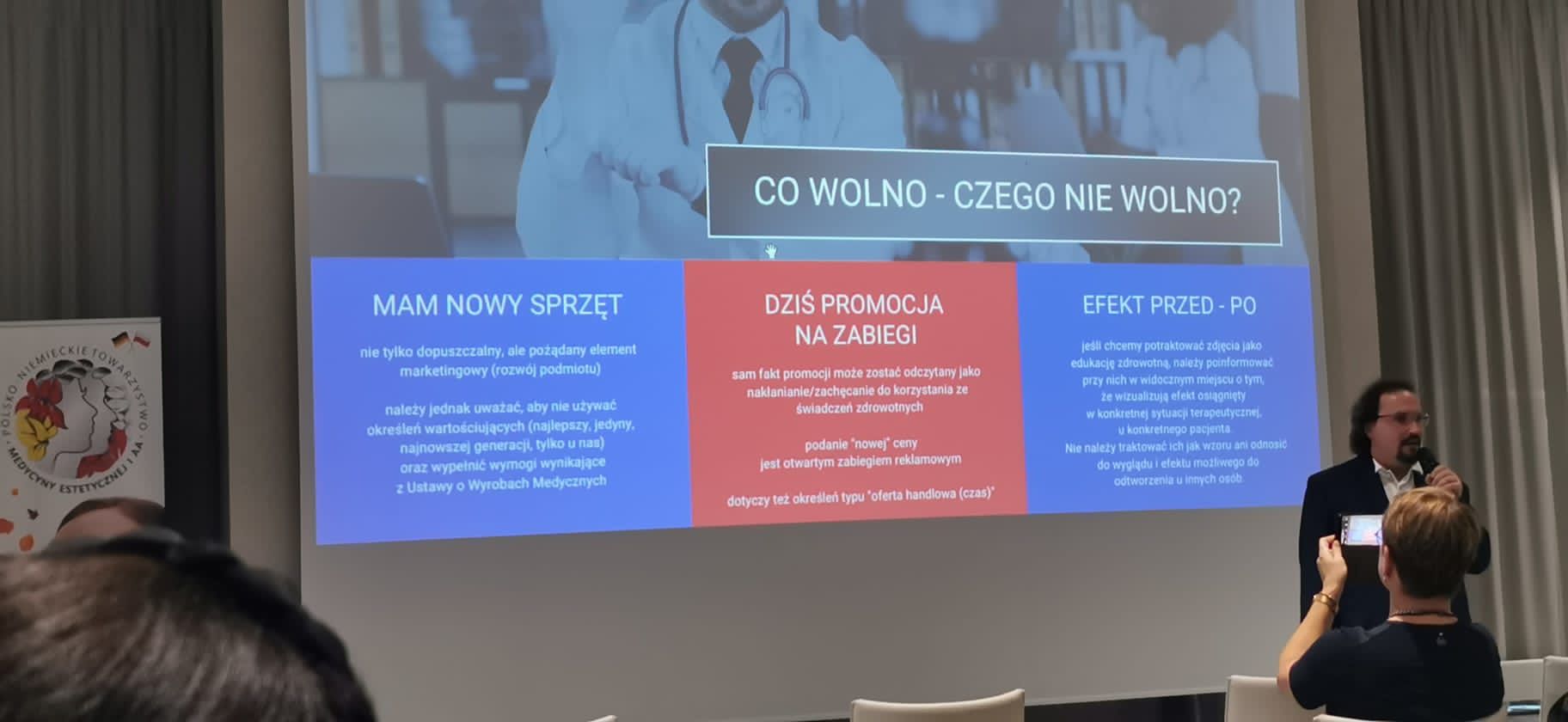 Zdjęcie przedstawiające mężczyznę wygłaszającego wykład, w tle prezentacja i słuchacze